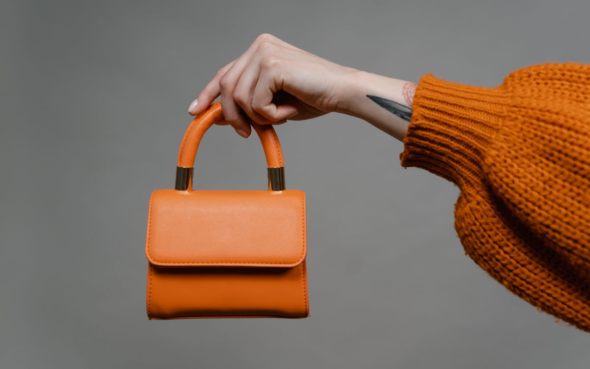 A person holding an orange leather shoulder bag at their side