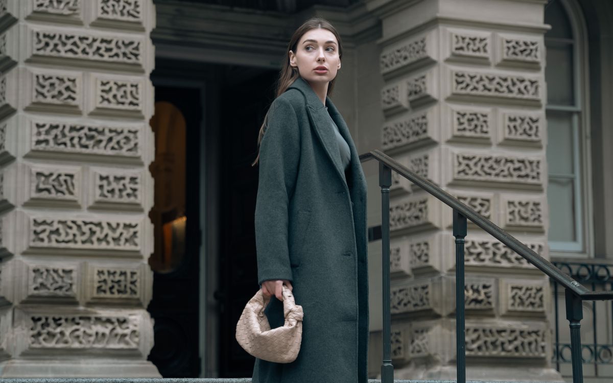 A model in a gray wool coat and palazzo pants carrying a beige handbag on steps